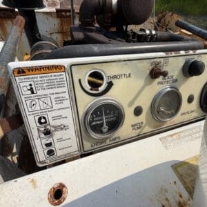 12 1 - Montana Auctions by Kevin Hill Control panel of a rusted industrial engine with a throttle gauge, stop switch, water pump gauge, and warning label visible outdoors.