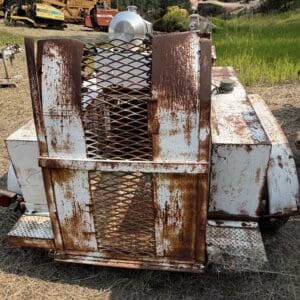 12 2 - Montana Auctions by Kevin Hill Rusty, white-painted utility trailer or equipment with a diamond-mesh metal grill, sitting on dry grass and dirt with other machinery in the background—aged and deteriorated metal surfaces visible.