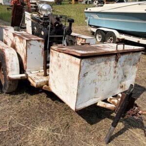 12 4 - Montana Auctions by Kevin Hill Rusty utility trailer with a large metal storage box and exposed engine, parked on dry grass near a boat trailer. The hitch and chains are visible in the foreground.