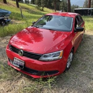 13 1 - Montana Auctions by Kevin Hill Red Volkswagen sedan parked on a grassy field with trees in the background and a trailer visible in the distance.