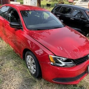 13 1 - Montana Auctions by Kevin Hill Red VW sedan parked on grassy ground, front hood dented with scratches, adjacent black SUV in background.