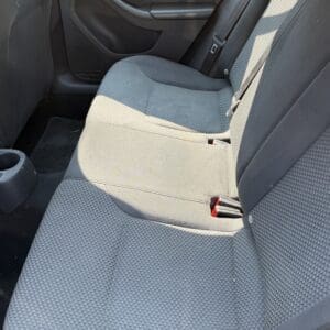 13 6 - Montana Auctions by Kevin Hill Backseat of a car with gray patterned cloth seats, door panel, and visible seat belt buckles in the middle and right positions, sunlight streaming in.