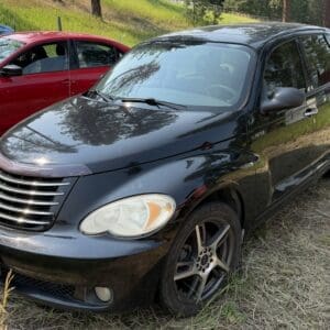14 2 - Montana Auctions by Kevin Hill Black Chrysler PT Cruiser parked on a grassy lot with a red car beside it; sunlight reflects off the hood and grille.