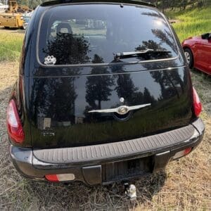 14 3 - Montana Auctions by Kevin Hill Rear view of a black Chrysler PT Cruiser with a tow hitch, parked on dry grass with a red car nearby and trees in the background.