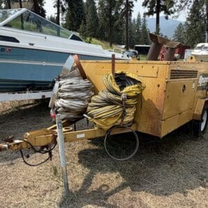 15 2 - Montana Auctions by Kevin Hill Yellow utility trailer with coiled cables and hoses, parked on dry grass near a blue boat in a wooded area.