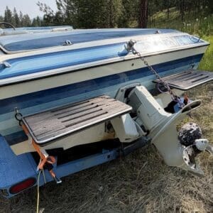 16 2 - Montana Auctions by Kevin Hill Blue-and-white speedboat on a trailer parked in dry grass; dual-prop outboard motor at the stern.