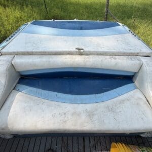 16 3 - Montana Auctions by Kevin Hill Back of a white and blue boat with dusty, weathered upholstery and padded seating outdoors on a trailer, grass and a fence in the background.
