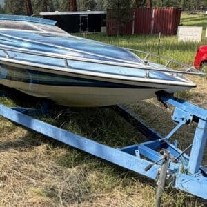 16 4 - Montana Auctions by Kevin Hill Blue and white speedboat on a blue trailer in a grassy field; a red car and storage sheds are in the background.
