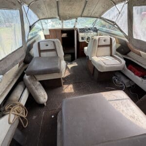 17 3 - Montana Auctions by Kevin Hill Old boat cockpit with beige seats, steering wheel, and dusty canopy in a cluttered cabin inside a small motorboat.