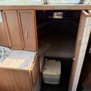 17 5 - Montana Auctions by Kevin Hill Wood-paneled boat cabin with open storage cabinets on the left and a small portable toilet in the doorway, berths with patterned upholstery beyond.