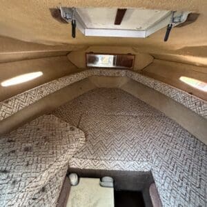 17 6 - Montana Auctions by Kevin Hill Interior of a small boat cabin with a V-shaped cushioned berth, beige patterned upholstery, and a small hatch/window at the far end.