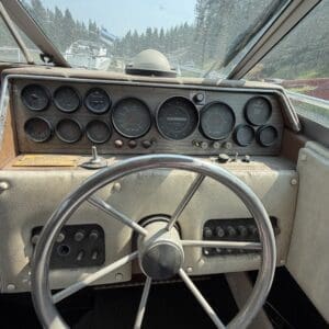 17 7 - Montana Auctions by Kevin Hill Vintage boat cockpit with a metal steering wheel and row of analog gauges on a dusty beige dashboard; exterior water and trees visible through the windshield.