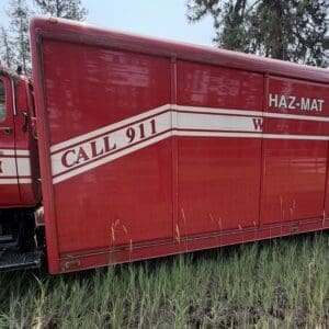 18 1 - Montana Auctions by Kevin Hill Red Haz-Mat Operations fire rescue truck with a 'CALL 911' diagonal stripe, parked on grass with trees in the background.