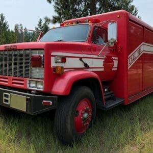 18 1 - Montana Auctions by Kevin Hill Red vintage fire/emergency rescue truck with GMC grille parked in a grassy field, white stripes along the side and CALL 911 text visible on the body.