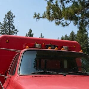 18 3 - Montana Auctions by Kevin Hill Front view of a red fire truck cab with flashing emergency lights against a blue sky and pine trees in the background.