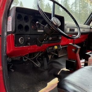 18 5 - Montana Auctions by Kevin Hill Interior of a red vintage truck showing a large black steering wheel and dashboard with round gauges and pedals visible under the dash outside trees. Accessory gear visible near center.