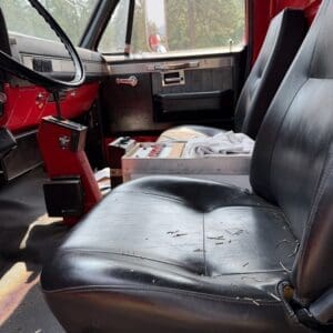 18 6 - Montana Auctions by Kevin Hill Interior of a red vintage truck cabin with a black leather seat scattered with pine needles and debris; steering wheel visible on the left.