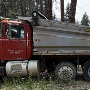 19 1 - Montana Auctions by Kevin Hill Red dump truck with a rusted bed parked on a dirt road in a forested area.
