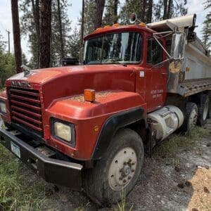 19 2 - Montana Auctions by Kevin Hill Red Mack dump truck parked on a dirt road in a pine forest, front grille and large bumper visible. The truck has a six-wheel configuration and a weathered, rugged look.