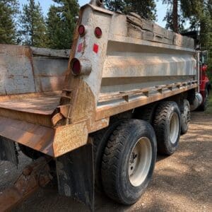 19 2 - Montana Auctions by Kevin Hill Rusty dump truck parked on a dirt road with three axles and large tires, forest in the background.