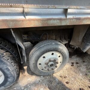 19 3 - Montana Auctions by Kevin Hill Rusty utility truck side with a flat tire, exposed wheel hub, and dusty undercarriage on a dirt surface with pine cones nearby.