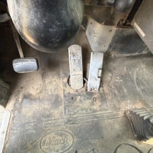 19 5 - Montana Auctions by Kevin Hill Dusty Mack truck cab interior with three pedals (clutch, brake, accelerator) and a worn rubber floor mat bearing the Mack logo.