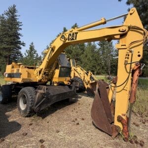 2 1 - Montana Auctions by Kevin Hill Yellow CAT excavator with a large arm and bucket parked on dry grass near pine trees in a rural lot.