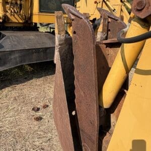 2 1 - Montana Auctions by Kevin Hill Close-up of a rusted excavator bucket attached to a yellow backhoe, with more heavy equipment in the background.