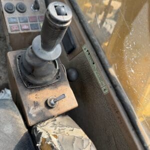 2 10 - Montana Auctions by Kevin Hill Industrial vehicle cockpit with a worn joystick and dusty controls, yellow exterior visible through a window.