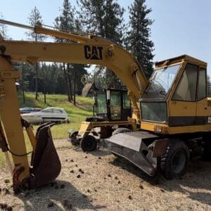 2 2 - Montana Auctions by Kevin Hill Yellow CAT excavator with a long boom and bucket resting on a grassy, pine‑tree backdrop.