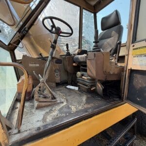 2 4 - Montana Auctions by Kevin Hill Dusty cab interior of a yellow construction excavator with a worn operator seat, steering wheel, and multiple levers.",