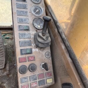2 9 - Montana Auctions by Kevin Hill Dusty operator panel in a yellow construction machine, featuring a joystick, three round gauges, and numerous worn buttons and switches inside the cab