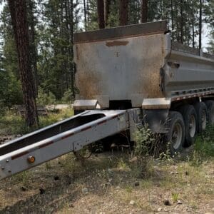 20 1 - Montana Auctions by Kevin Hill Rusty dump truck trailer parked in a forested area with tall trees and overgrown ground, showing a long loading ramp.