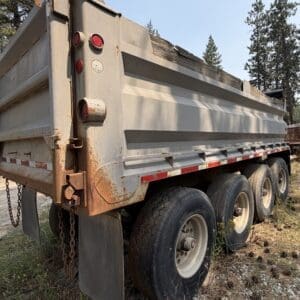 20 2 - Montana Auctions by Kevin Hill Rusty dump trailer with red lights, chains, and weathered wood on top, parked in a forested area with tall trees nearby.