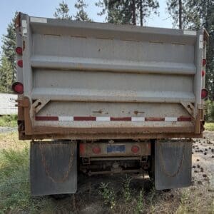 20 3 - Montana Auctions by Kevin Hill Rear view of a dirty, rusted dump truck with mud flaps, chains, and red rear lights in a grassy lot.