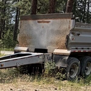 20 5 - Montana Auctions by Kevin Hill Rusty dump trailer with six wheels parked on a dirt road in a pine forest.