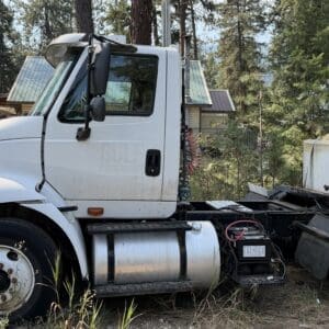 21 1 - Montana Auctions by Kevin Hill White semi-truck tractor with no trailer sits in an overgrown yard among pine trees and solar panels in the background.
