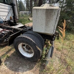 21 4 - Montana Auctions by Kevin Hill Truck rear wheel with a large concrete block loaded on a frame, in a rural, overgrown lot with pine trees nearby