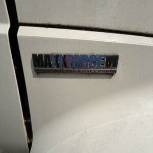 22 3 - Montana Auctions by Kevin Hill Chrome badge on a white vehicle reading 'MAXFORCED' with smaller text 'INTERNATIONAL DIESEL POWER' beneath, weathered with dirt.