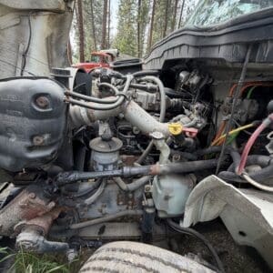 22 4 - Montana Auctions by Kevin Hill Close-up of a rusted truck engine with exposed hoses and wiring, a dusty front wheel in the foreground and trees in the background, indicating outdoor exposure.