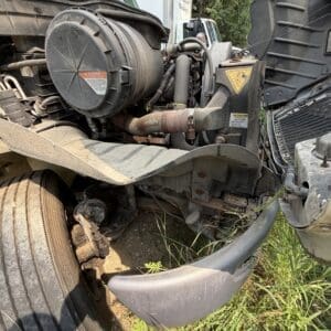 22 6 - Montana Auctions by Kevin Hill Close-up of a heavy truck's front end, exposing the engine bay, hoses, radiator, and a large dusty tire with grass beneath.