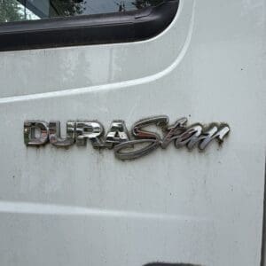 22 7 - Montana Auctions by Kevin Hill Chrome DURASTAR badge on a white vehicle door, close-up view showing dirt and partial window edge.