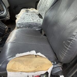 22 8 - Montana Auctions by Kevin Hill Front driver seat of a truck with torn black seat cover exposing yellow foam and damaged upholstery, dashboard and window visible.",
