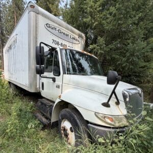 23 1 - Montana Auctions by Kevin Hill Front three-quarter view of a white box truck parked in tall grass beside trees, overgrown vegetation surrounding the vehicle.