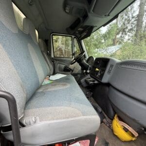 23 1 - Montana Auctions by Kevin Hill Interior of a work truck cab with a long gray-blue bench seat, steering wheel, and clutter under the seat, including a yellow hard hat and red tools on the floor outside the pedals.