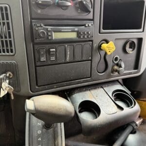 23 5 - Montana Auctions by Kevin Hill Dashboard of a vehicle with climate controls, radio, and multiple switches; yellow parking brake knob, cup holders, and a gear selector showing P R N D 2 1 with keys in the ignition on the left.