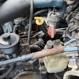 23 7 - Montana Auctions by Kevin Hill Close-up of a dusty vehicle engine compartment showing hoses, wiring, and a rusty pipe crossing in front of a translucent coolant reservoir with yellow and red valve handles nearby