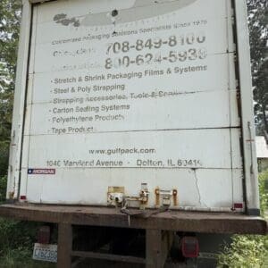 23 9 - Montana Auctions by Kevin Hill Rear of a weathered white truck trailer with faded company text and two phone numbers, rusted bumper, and surrounding vegetation empty lot nearby.