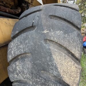 24 2 - Montana Auctions by Kevin Hill Close-up of a large dusty tractor tire with deep tread leans against a yellow construction machine outdoors, with grass and trees in the background.