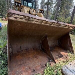 24 2 - Montana Auctions by Kevin Hill Rusty bulldozer bucket resting on the ground in a pine forest.